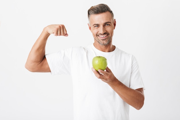 Retrato de um homem europeu dos anos 30 com cerdas vestindo camiseta casual mostrando seus bíceps e segurando uma maçã isolada no branco