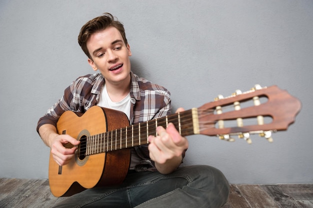 Retrato de um homem feliz sentado no chão tocando violão na parede cinza