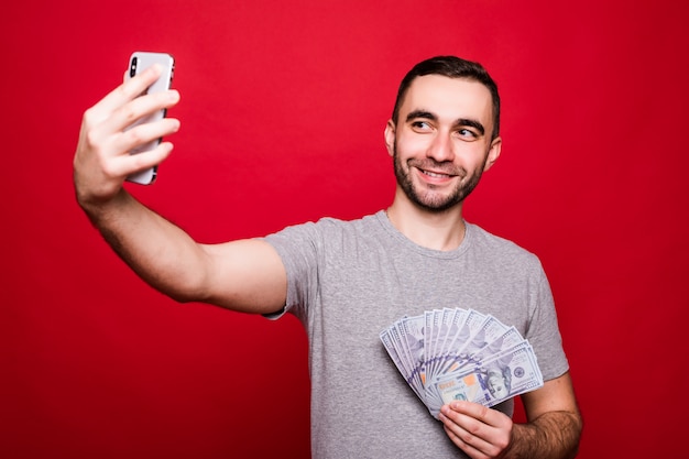 Retrato de um homem feliz tirando uma selfie com o celular, mostrando notas de dinheiro em pé sobre fundo vermelho