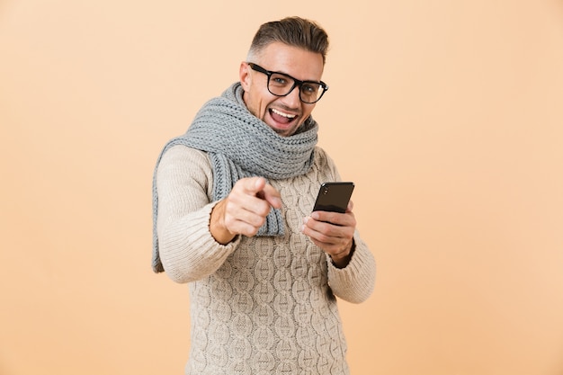 Retrato de um homem feliz, vestido com um suéter e um lenço, em pé, isolado sobre uma parede bege, segurando um telefone celular