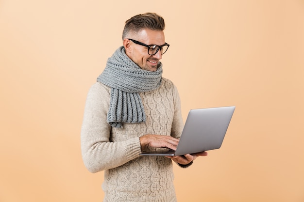 Retrato de um homem feliz, vestido com um suéter e um lenço, em pé, isolado sobre uma parede bege, trabalhando em um computador laptop