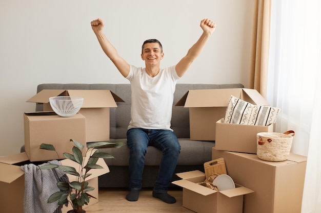 Retrato de um homem feliz, vestindo camiseta branca e jeans, sentado no sofá cercado de caixas de papelão, braços levantados, comemorando a mudança, expressando emoções positivas.