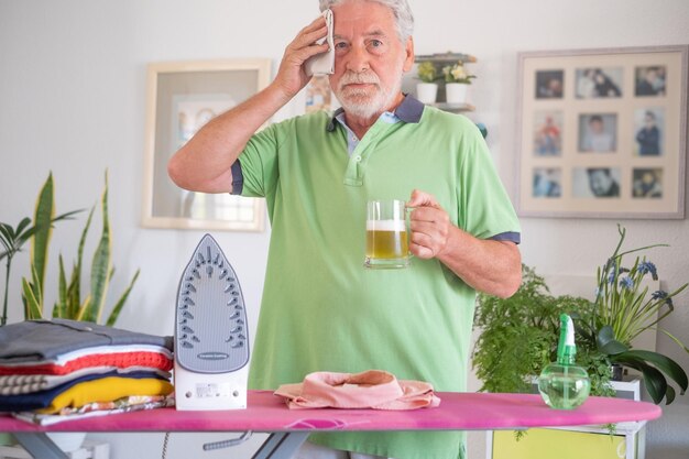 Retrato de um homem idoso ajudando nas tarefas domésticas passando roupas limpa a testa cansada do trabalho enquanto segura um copo de cerveja na mão