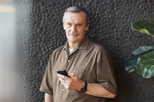 Retrato de um homem idoso e sorridente, caucasiano, com uma camisa marrom e um relógio de pulso em pé contra um fundo de textura de pedra e usando o telefone