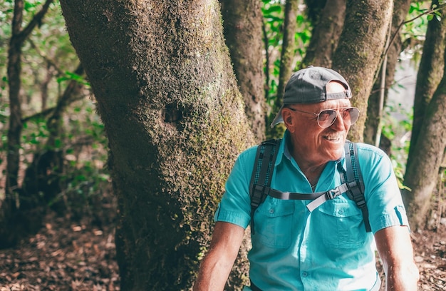 Retrato de um homem idoso sorridente com óculos de sol e mochila sentado em um dia de caminhada na floresta