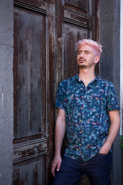 Retrato de um homem jovem turista com camisa azul verão e cabelo rosa pastel, inclinando-se ao lado de uma porta de madeira escura.