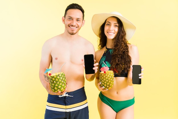 Retrato de um homem latino atraente e mulher caucasiana mostrando sua tela de smartphone e sorrindo enquanto segura um coquetel de pina colada