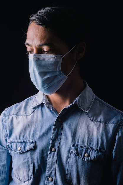 Retrato de um homem latino olhando para baixo desanimado usando máscara médica e camisa azul em fundo preto. Efeitos do coronavírus