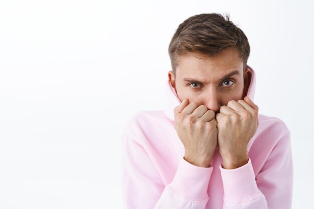 Foto retrato de um homem loiro bonito com um capuz rosa, sentindo frio ou deprimido, ficando em casa se enrolando em roupas, parecendo ofendido ou inseguro na frente por baixo da testa