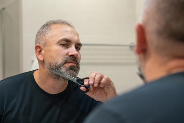 Foto retrato de um homem maduro bonito aparando a própria barba em pé no banheiro