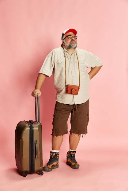 Retrato de um homem maduro e obeso em roupas casuais com mala e câmera vintage posando sobre fundo de estúdio rosa Turista