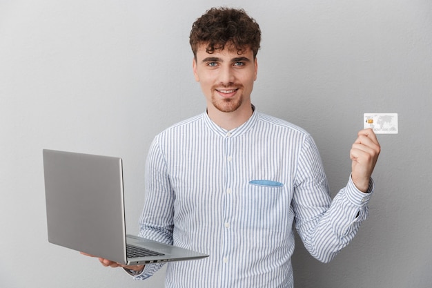 Retrato de um homem masculino vestido com uma camisa, sorrindo enquanto segura um laptop prateado e um cartão de crédito isolado sobre uma parede cinza