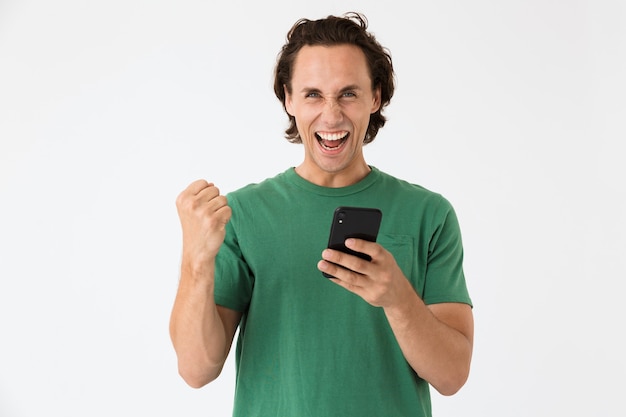 Retrato de um homem moreno encantado, vestindo uma camiseta básica, gritando e segurando o smartphone isolado sobre uma parede branca
