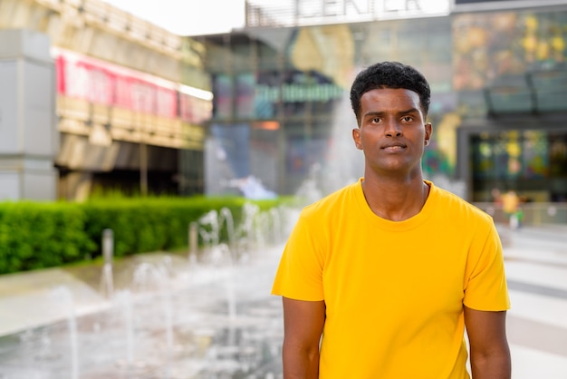 Retrato de um homem negro africano bonito vestindo uma camiseta amarela ao ar livre na cidade durante o verão ao lado da fonte