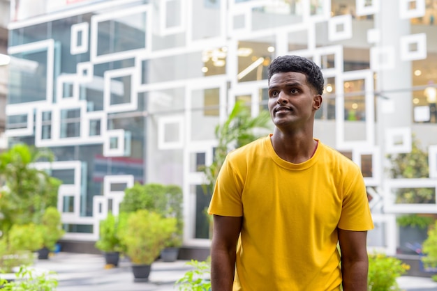 Retrato de um homem negro africano bonito vestindo uma camiseta amarela ao ar livre na cidade durante o verão enquanto pensa