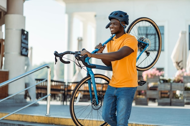 Retrato de um homem nigeriano sorridente e positivo segurando uma bicicleta usando um capacete de segurança na rua urbana