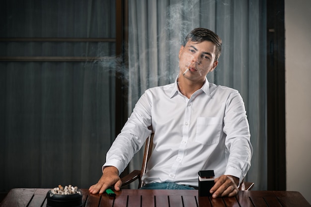 Retrato de um homem posando enquanto está sentado em uma mesa em que está um cinzeiro cheio de cigarros