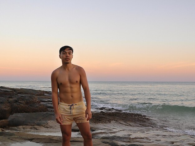 Foto retrato de um homem sem camisa de pé na praia contra o céu claro durante o pôr do sol