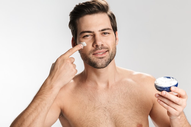 Retrato de um homem seminu satisfeito olhando para a câmera e aplicando creme facial isolado no branco