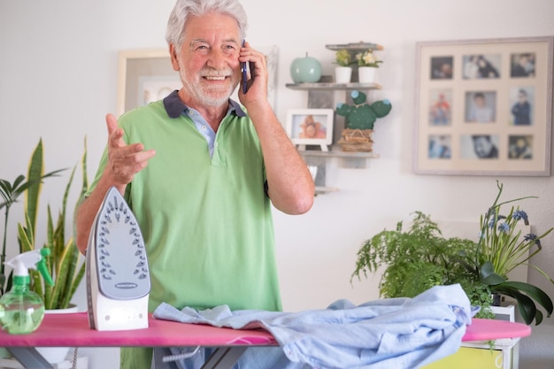 Retrato de um homem sênior alegre em casa ajudando com o trabalho doméstico passando camisas enquanto usa seu telefone celular