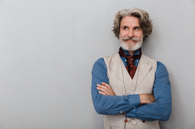 Retrato de um homem sênior otimista satisfeito barbudo com cabelos grisalhos isolado sobre o fundo da parede cinza.