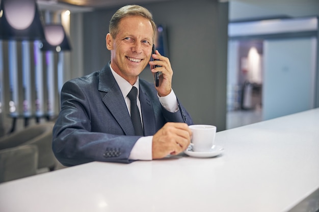 Retrato de um homem sorridente de terno elegante e gravata tomando café e falando no celular no bar