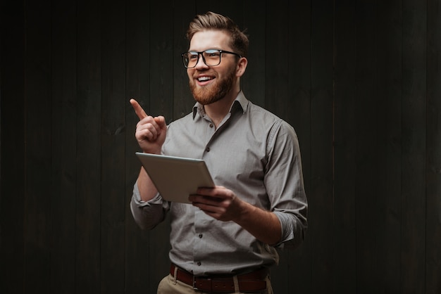 Foto retrato de um homem sorridente e feliz usando óculos, segurando um computador tablet e apontando o dedo, isolado em uma superfície de madeira preta