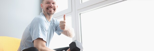 Retrato de um homem sorridente segurando halteres e mostrando o gesto de polegar para cima