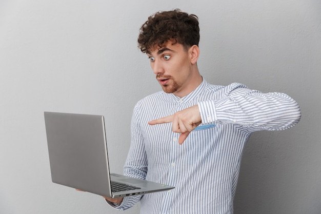 Retrato de um homem surpreso, vestido com uma camisa, pensando enquanto segura e aponta para um laptop prateado isolado sobre uma parede cinza