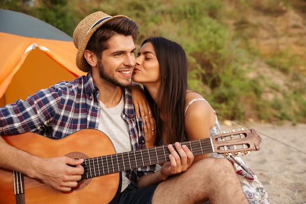 Retrato de um homem tocando violão para a namorada sentado em uma barraca de acampamento na praia