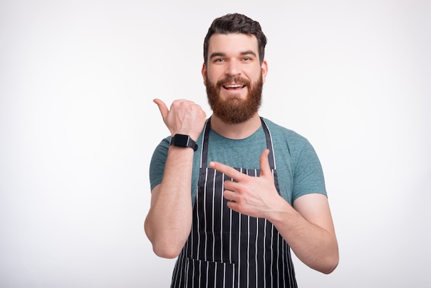 Retrato de um homem vestindo um avental de cozinha na parede branca está apontando para o seu smartwatch