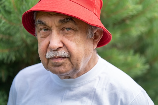 Retrato de um idoso de 70 anos de camiseta branca e chapéu vermelho que olha com calma e atenção para a câmera, no jardim do campo.