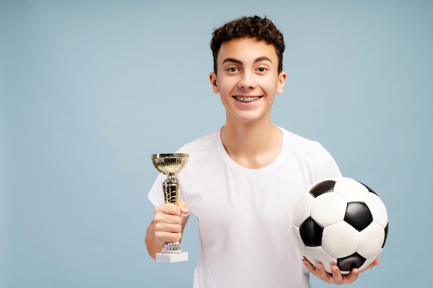 Retrato de um jogador de futebol adolescente entusiasmado com aparelhos dentários segurando uma taça de troféu e uma bola de futebal