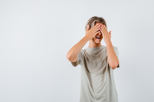 Retrato de um jovem adolescente cobrindo os olhos com as mãos na camiseta e olhando animado para a frente