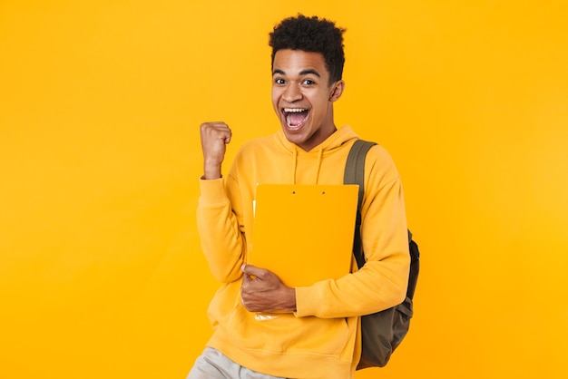 Retrato de um jovem adolescente feliz em pé isolado na parede amarela, carregando mochila e livros didáticos, comemorando o sucesso