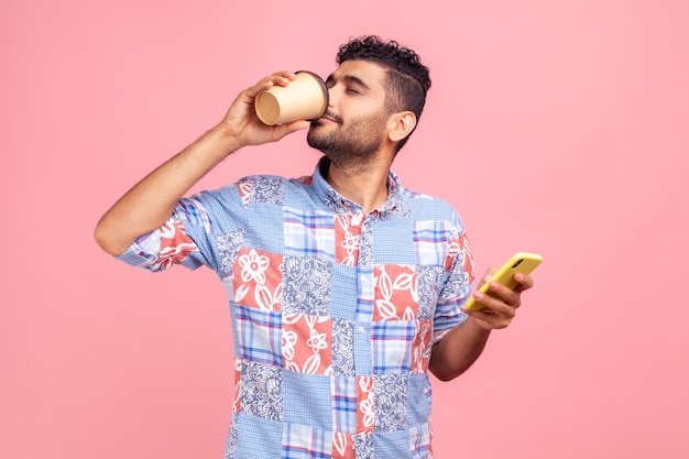 Retrato de um jovem adulto atraente na camisa de estilo casual em pé e segurando o copo descartável e telefone bebendo e desfrutando de bebida quente Foto de estúdio interior isolada no fundo rosa