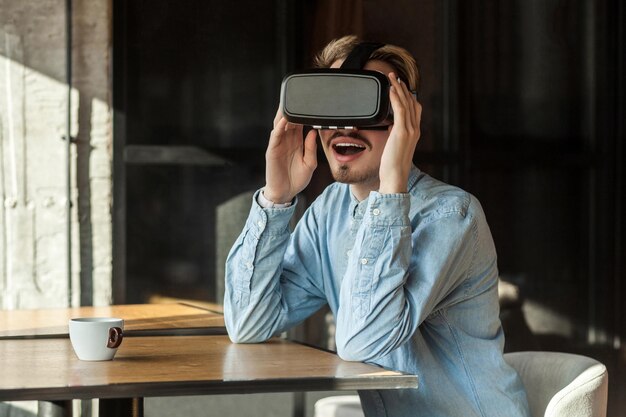 Retrato de um jovem adulto barbudo inacreditável feliz na camisa jeans azul sentado no café usando vr e assistindo vídeo no simulador com cara feliz incrível Estilo de vida interior