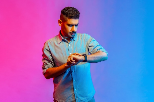 Retrato de um jovem adulto concentrado na camisa em pé olhando para seu smartwatch verificando o tempo sendo apressado para conhecer o estúdio interno isolado no fundo colorido da luz de néon