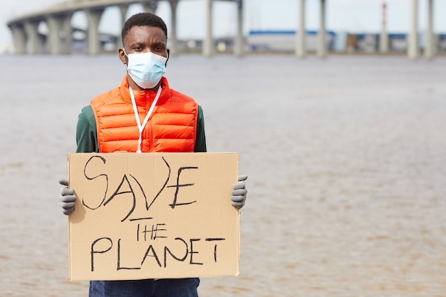 Retrato de um jovem africano com uma máscara protetora segurando um cartaz em pé perto da costa