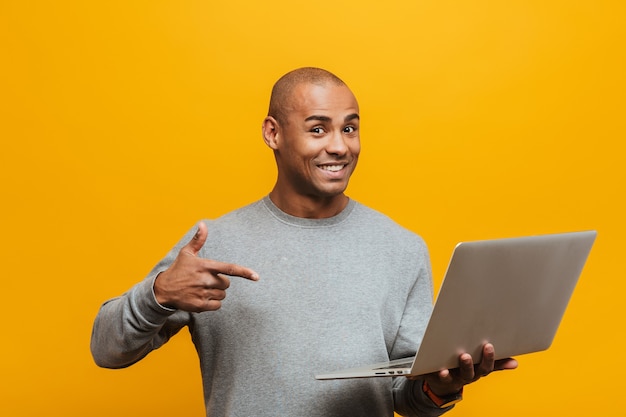 Retrato de um jovem africano confiante e sorridente atraente em pé sobre a parede amarela, apontando para o computador portátil