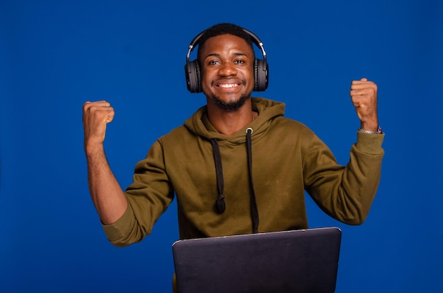 Retrato de um jovem africano feliz ouvindo música com fones de ouvido em pé e usando laptop