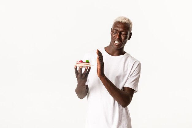 Retrato de um jovem afro-americano bonito enojado, não querendo comer sobremesa, se encolhendo com um bolo nojento