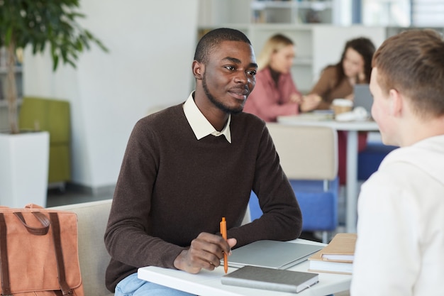 Retrato de um jovem afro-americano conversando com um amigo do outro lado da mesa enquanto estudava na biblioteca da faculdade,