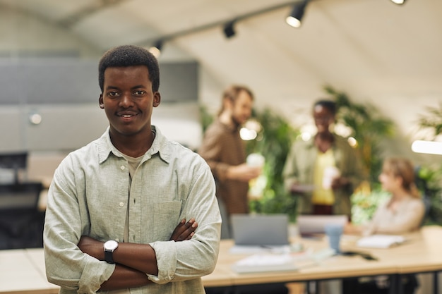 Retrato de um jovem afro-americano da cintura para cima enquanto posa com confiança em pé com os braços cruzados em um escritório moderno com pessoas trabalhando na superfície, copie o espaço