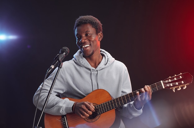 Retrato de um jovem afro-americano tocando guitarra no palco e cantando ao microfone sob as luzes da cintura para cima, copie o espaço