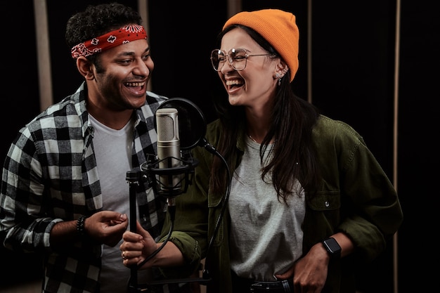 Retrato de um jovem alegre e uma mulher em dueto cantando em um microfone condensador durante a gravação de um