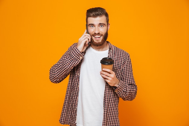 Retrato de um jovem alegre, vestindo roupas casuais, falando no celular e bebendo café para viagem