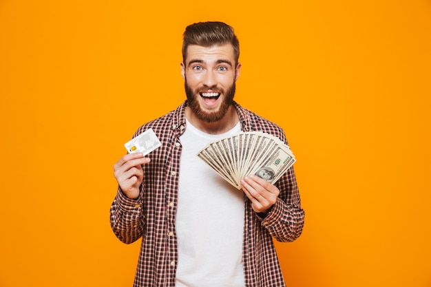 Retrato de um jovem alegre, vestindo roupas casuais, segurando notas de dinheiro e mostrando um cartão de crédito de plástico