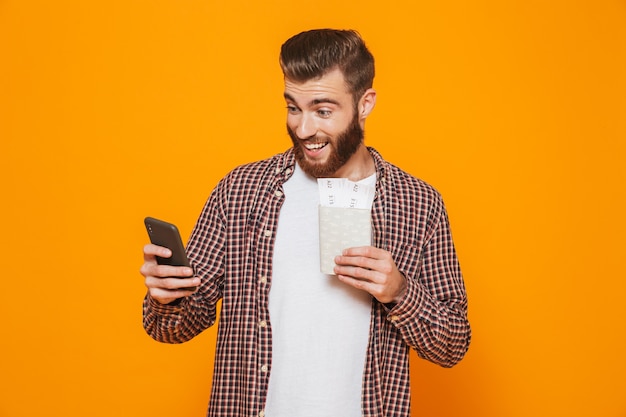 Retrato de um jovem alegre, vestindo roupas casuais, segurando o passaporte, usando telefone celular