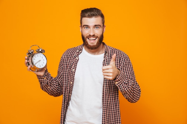 Retrato de um jovem alegre, vestindo roupas casuais, segurando um despertador mostrando os polegares para cima